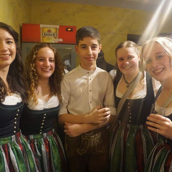 Fünf Personen posieren für ein Foto, vier in traditioneller Kleidung und eine in Weiß. Ein Kühlschrank mit Kaiser-Bier ist im Hintergrund.