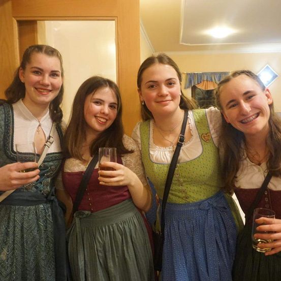Vier Frauen in traditioneller deutscher Kleidung lächeln und posieren für ein Foto, jede hält ein Glas mit Getränk. Sie stehen eng beieinander, mit einer Holztür und einem Spiegel im Hintergrund.