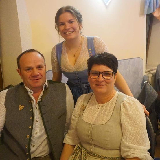 Drei Personen posieren für ein Foto in einem Raum. Der Mann vorne lächelt. Die Frau hinter ihm hat ihre Hand auf der Schulter der Frau vorne. Die Frau vorne trägt eine Brille.