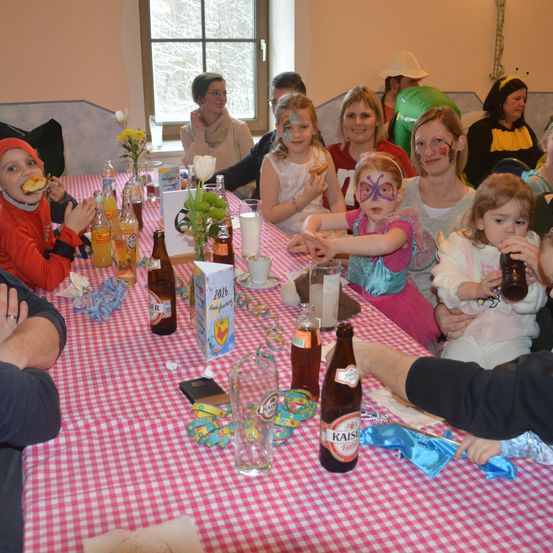 Eine Gruppe von Menschen, einschließlich Kindern, sitzt um einen Tisch mit Getränken, Essen und Dekorationen. Einige sind in Kostümen gekleidet und haben Gesichtsbemalung.