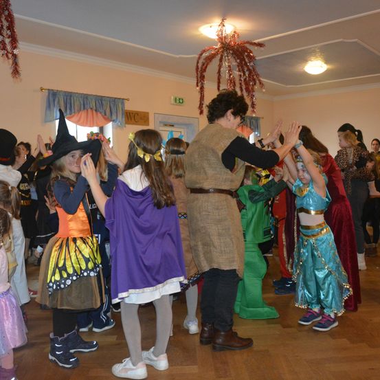 Eine Gruppe von Kindern in verschiedenen Kostümen, einschließlich einer Hexe und einer Fee, tanzt in einem Raum. Eine erwachsene Frau in der Mitte, die einen Gürtel trägt, führt die Kinder an.