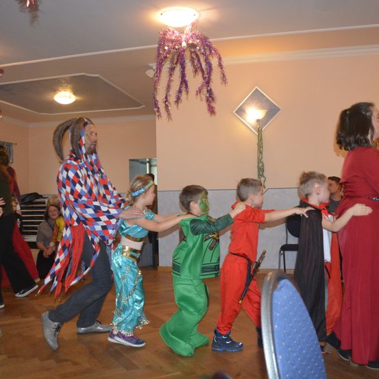 Eine Gruppe von Kindern und Erwachsenen in Kostümen tanzt in einem Raum, ein Mann mit Clownsmaske hält ein Mädchen und zwei Jungen in grünen und roten Kostümen.