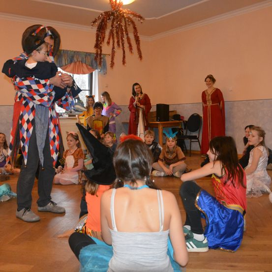 Kinder in Kostümen sitzen im Kreis und beobachten einen Mann in Clown-Kostüm, der ein Kind hält. Zwei Frauen in roten Kleidern stehen auf der Bühne mit Mikrofon und Lautsprecher.