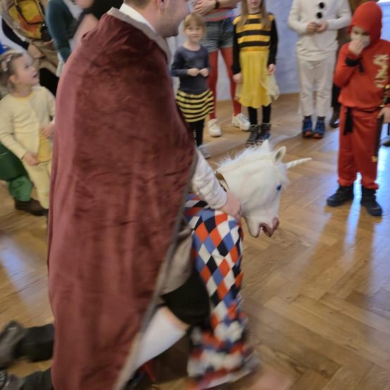 Ein Mann in einem Gewand hält ein Einhorn-Spielzeug, während er auf einem Boden sitzt. Mehrere Kinder beobachten und sind in verschiedenen Kostümen gekleidet.