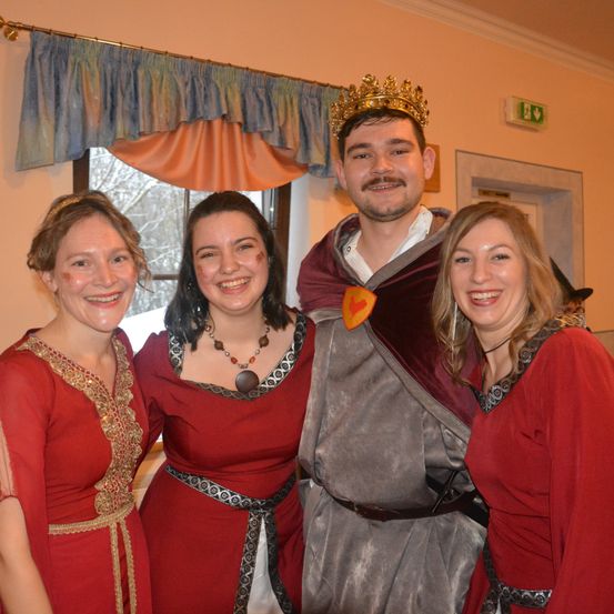 Vier Menschen, gekleidet in mittelalterliche Kostüme, lächeln und posieren für ein Foto. Der Mann in der Mitte trägt eine Krone und einen Umhang. Die drei Frauen an den Seiten tragen rote Kleider mit goldenen Verzierungen. Dahinter befindet sich ein Fenster mit einem Vorhang.