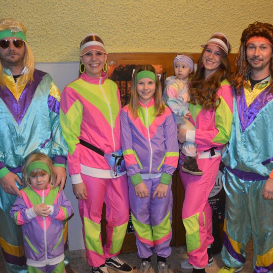 Eine Familie, gekleidet in leuchtenden, retro 80er-Jahre-Trainingsanzüge, posiert drinnen. Erwachsene und Kinder lächeln für die Kamera und zeigen ihre bunten Kostüme.
