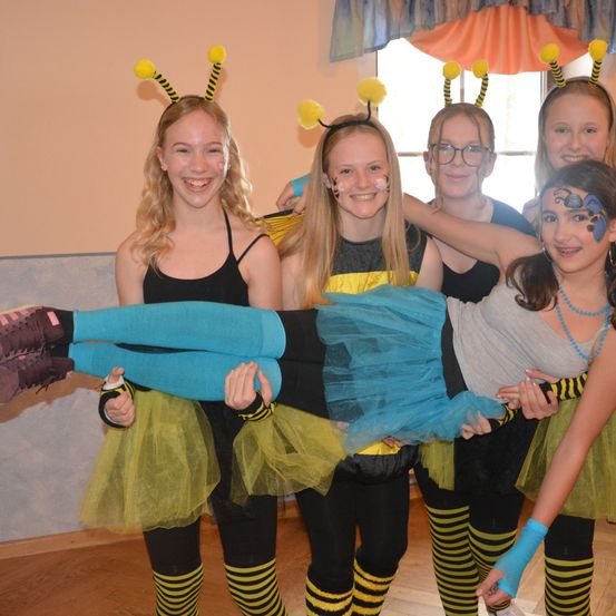 Eine Gruppe junger Mädchen, die Bienenkostüme tragen, hält ein Mädchen auf ihren Schultern, während sie für ein Foto in einem Raum mit Holzboden posiert.