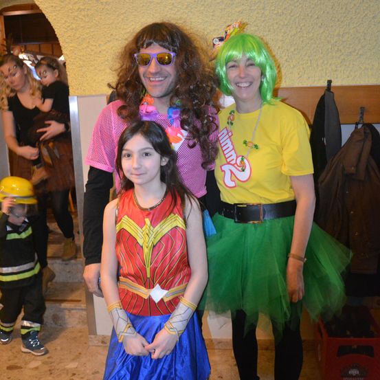 Eine Gruppe von Menschen in Kostümen, ein Mann mit Perücke, eine Frau in einem Tutu, ein Mädchen in einem Wonder-Woman-Kostüm und ein Kind in einem Feuerwehrmannkostüm, stehen drinnen.