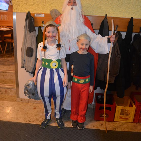 Zwei Kinder in Kostümen stehen neben einem Mann, der als Weihnachtsmann gekleidet ist. Der Mann hält einen Holzstock und trägt einen roten Umhang. Hinter ihnen hängen Kleidungsstücke an einem Kleiderständer.