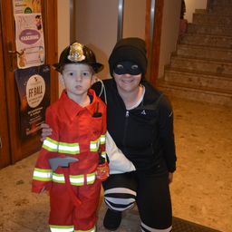 Ein Junge in einem Feuerwehrmann-Kostüm posiert mit einer vermummten Frau. Sie stehen neben einer Tür mit einem Plakat für Kinderfasching.