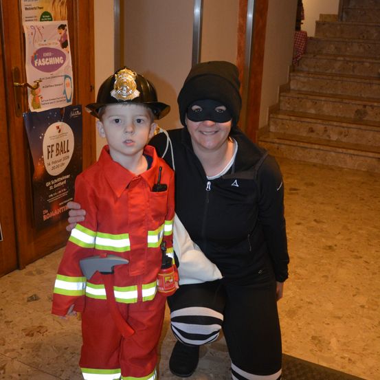 Ein Junge in einem Feuerwehrmann-Kostüm posiert mit einer vermummten Frau. Sie stehen neben einer Tür mit einem Plakat für Kinderfasching.