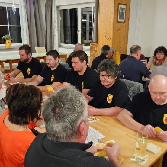 Eine Gruppe von Erwachsenen, einige in schwarzen T-Shirts, sitzen um einen Tisch herum, möglicherweise in einer Besprechung oder Zusammenkunft. Sie halten Spielkarten und scheinen an einem Spiel teilzunehmen.