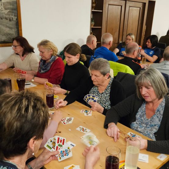 Mehrere ältere Erwachsene sitzen um einen Tisch und spielen ein Kartenspiel. Einige tragen Brillen und einige haben Getränke vor sich. An der Wand hinter ihnen hängt ein Bild.