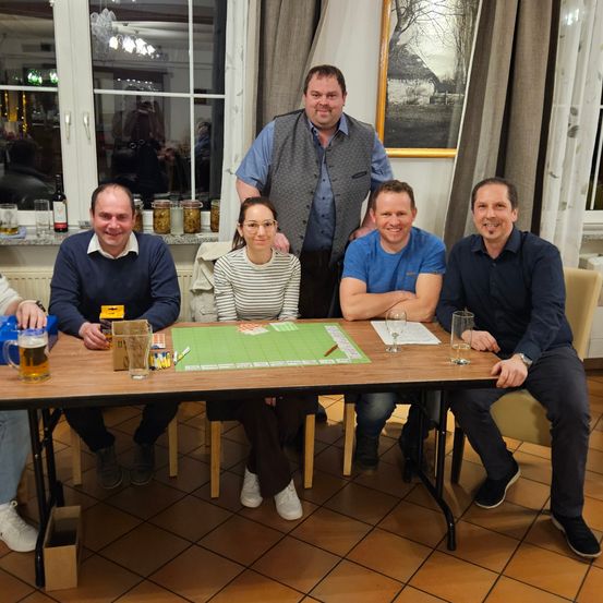 Fünf Personen sitzen um einen Tisch mit Spielbrett und Getränken. Drei Männer lächeln, während eine Frau in die Kamera schaut. Dahinter steht ein Mann mit verschränkten Armen.
