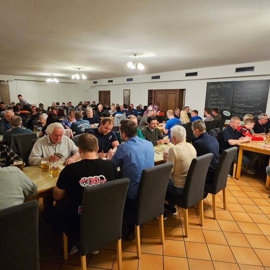 Eine große Gruppe von Menschen sitzt in einem Speisesaal um Tische herum und beschäftigt sich mit verschiedenen Aktivitäten. Einige essen, andere spielen Karten. Der Raum hat eine Tafel mit Schriftzügen und der Boden ist gefliest.