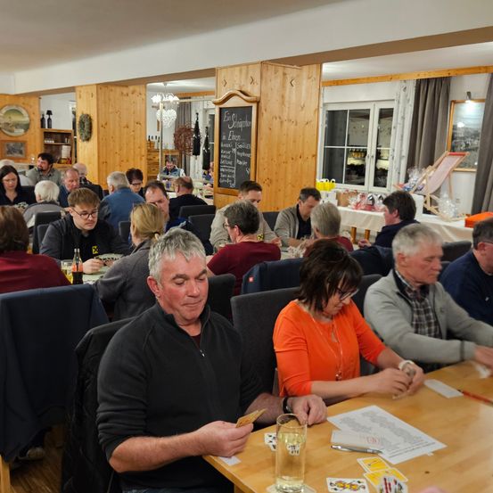 Eine Gruppe von Menschen sitzt in einem Restaurant um einen Tisch herum, einige spielen Karten. Ein Mann vorn schaut auf eine Karte in seiner Hand.