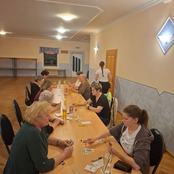 Eine Gruppe von Frauen sitzt um einen langen Tisch herum und spielt Karten. Eine Frau steht im Hintergrund. Auf dem Tisch befinden sich Gläser, Karten und Papiere.