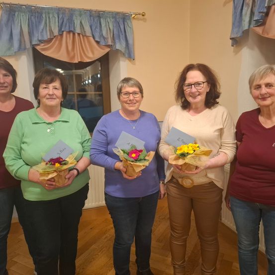 Fünf Frauen mit Blumen und Karten stehen in einem Zimmer mit Fenster und Holzboden.
