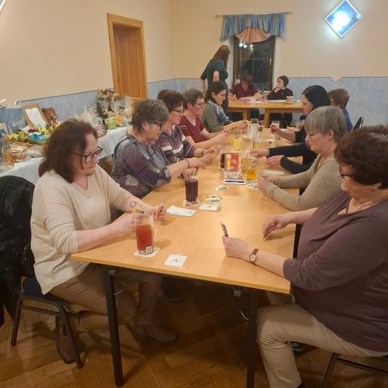 Eine Gruppe von Frauen sitzt in einem Raum um einen Tisch herum, trinkt und unterhält sich. Ein Tisch mit Essen steht links.