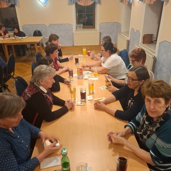 Eine Gruppe älterer Frauen sitzt um einen Tisch herum, wahrscheinlich in einem sozialen Club. Gläser und Karten sind auf dem Tisch. Einige Frauen tragen Brillen und Halsketten. Ein Fenster und ein Heizkörper sind im Hintergrund.