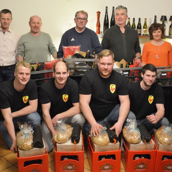 Eine Gruppe von Männern und Frauen in schwarzen T-Shirts mit Logos posiert für ein Foto. Sie halten Brot in Plastiktüten und stehen hinter roten Boxen. Flaschen stehen auf einem Regal hinter ihnen.