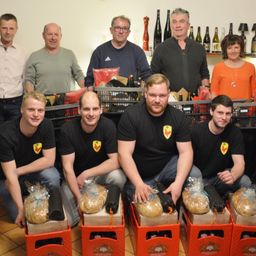 Eine Gruppe von Männern und Frauen in schwarzen T-Shirts mit Logos posiert für ein Foto. Sie halten Brot in Plastiktüten und stehen hinter roten Boxen. Flaschen stehen auf einem Regal hinter ihnen.