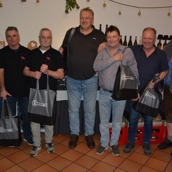 Fünf Männer stehen nebeneinander und halten Einkaufstüten. Sie tragen alle Freizeitkleidung. Dahinter steht ein Tisch mit Weinflaschen und ein Regal mit Lampen. Der Boden ist gefliest.