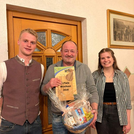 Drei Personen stehen vor einer Tür. Der Mann in der Mitte hält ein Zertifikat und einen Korb mit Essen. Der Mann links trägt eine Weste, und die Frau rechts trägt ein kariertes Hemd. Hinter ihnen ist ein Bilderrahmen.