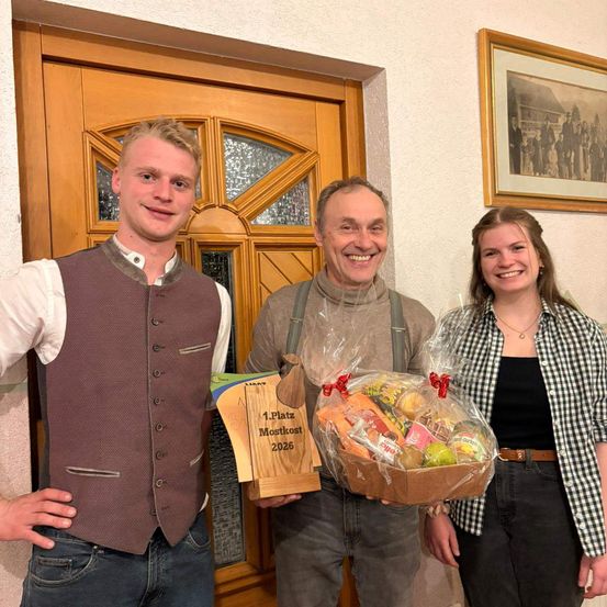 Drei Personen stehen vor einer Tür. Der Mann auf der linken Seite hält eine Plakette für den 1. Platz Mostkost 2026. Der Mann in der Mitte hält einen Korb mit Lebensmitteln. Die Frau auf der rechten Seite lächelt.
