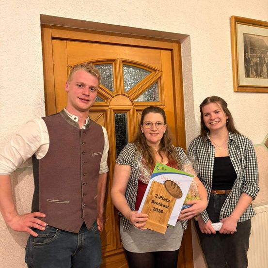 Drei Personen stehen vor einer Holztür. Die Frau in der Mitte hält eine Schachtel und ein Büchlein. Der Mann auf der linken Seite trägt eine Weste und Jeans. Die Frau auf der rechten Seite trägt eine Halskette und ein kariertes Hemd.