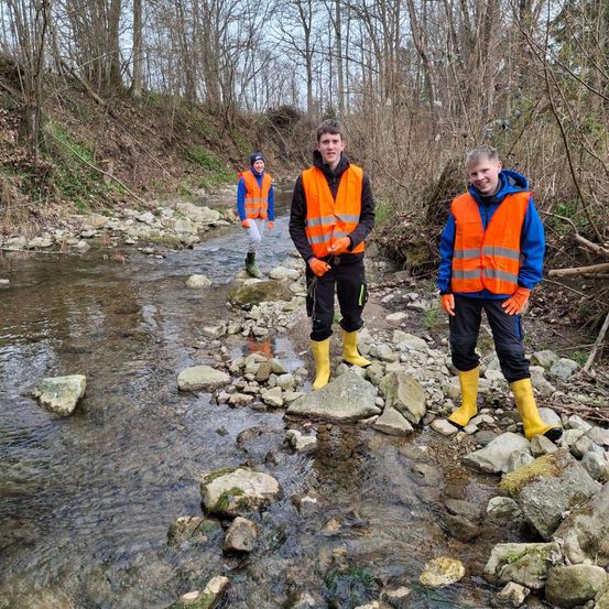 Drei Personen in orange Warnwesten und gelben Stiefeln stehen auf Felsen in der Nähe eines Baches im Wald.