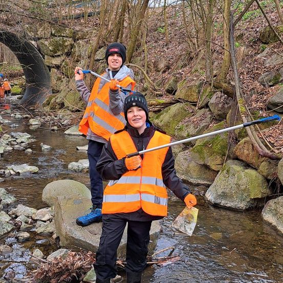 Zwei Kinder in orangefarbenen Westen stehen in einem Bach und halten einen langen Stock. Hinter ihnen ist eine Brücke über dem Wasser zu sehen.
