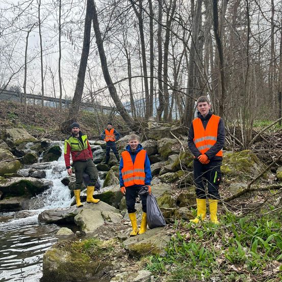 Vier Personen in Warnwesten und Gummistiefeln stehen auf Felsen am Bach. Einer hält einen Müllsack. Bäume und eine Brücke im Hintergrund.