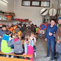 Eine Gruppe von Menschen sitzt an Tischen in einem Raum. Eine Frau in einer blauen Jacke hält ein Glas Bier. Eine andere Frau in einer braunen Jacke lächelt. Einige Verkehrszeichen hängen an der Wand.