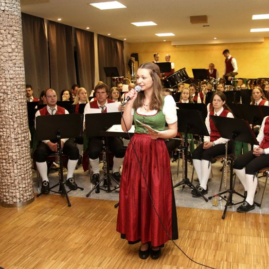 Eine Frau in traditioneller deutscher Kleidung steht vor einer Band und hält ein Mikrofon. Die Band sitzt mit verschiedenen Instrumenten, während einige Mitglieder im Hintergrund stehen.