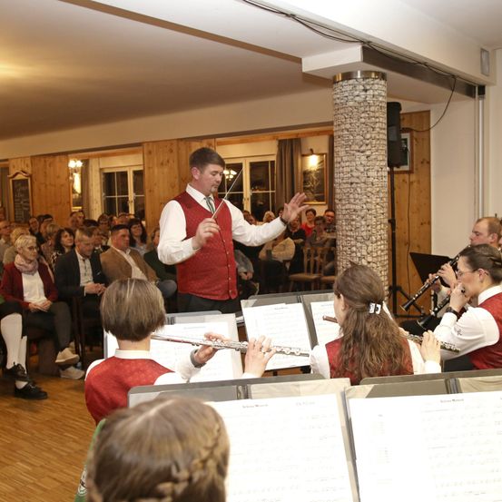 Ein Dirigent steht vor einem Publikum und leitet eine Aufführung. Mehrere Musiker spielen Instrumente wie Flöten und Klarinetten, die vor Notenständern sitzen. Die Umgebung erscheint feierlich, mit hölzernen Wänden und einer Kronleuchter an der Decke.