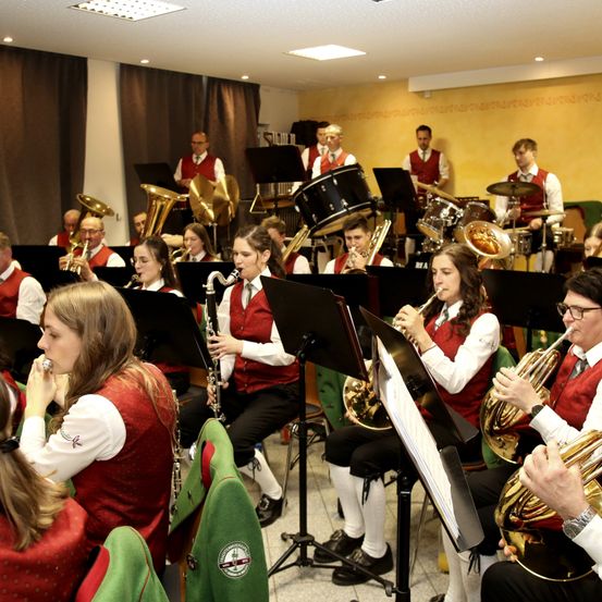 Eine Blechbläsergruppe spielt in einem Raum mit gelben Wänden und trägt rote Westen und weiße Hemden. Sie spielen Instrumente wie Trompeten, Posaunen und Saxophon.