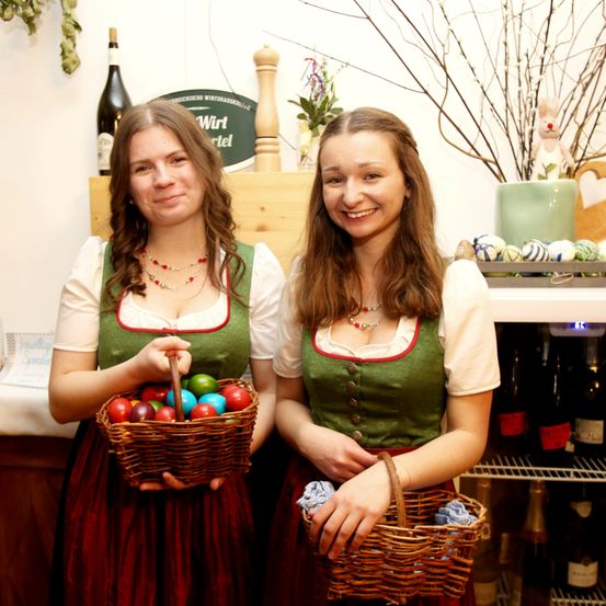 Zwei Frauen in traditioneller grüner Kleidung halten Körbe mit bunten Eiern und lächeln für ein Foto. Ein Kühlschrank mit Weinflaschen und ein Regal mit Osterdekoration sind im Hintergrund zu sehen.