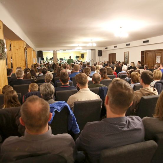 Eine Menschenmenge sitzt in Stühlen in einem Raum, wahrscheinlich bei einem Seminar. Einige Leute schauen auf die Bühne, wo zwei Personen stehen.