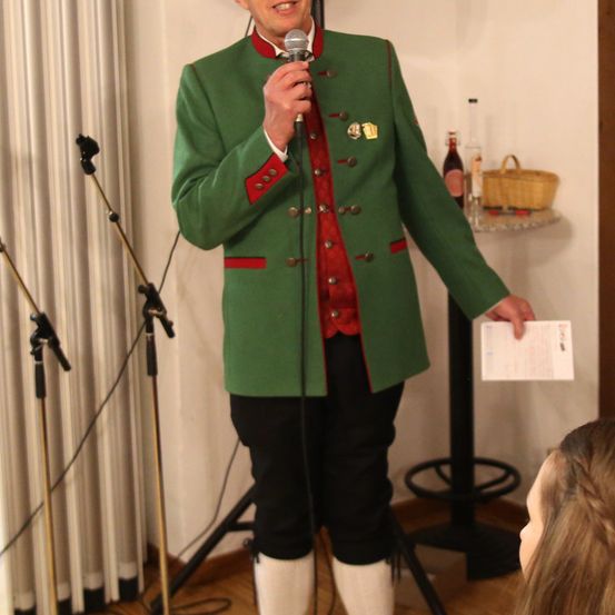 Ein Mann in einer grünen und roten Jacke hält ein Mikrofon. Er lächelt und scheint eine Rede zu halten. Hinter ihm steht ein Tisch mit einem Korb und einer Flasche.