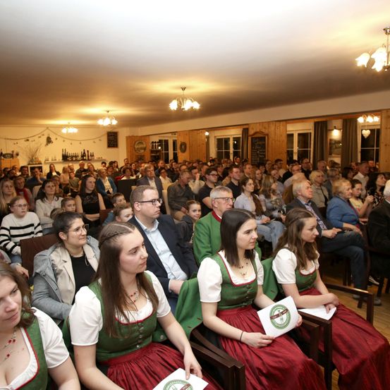 Eine große Menschenmenge versammelt sich in einem Raum, wahrscheinlich einem Konferenzsaal. Einige tragen traditionelle Kostüme, und die meisten schauen auf einen Mann vorne. Andere unterhalten sich miteinander.