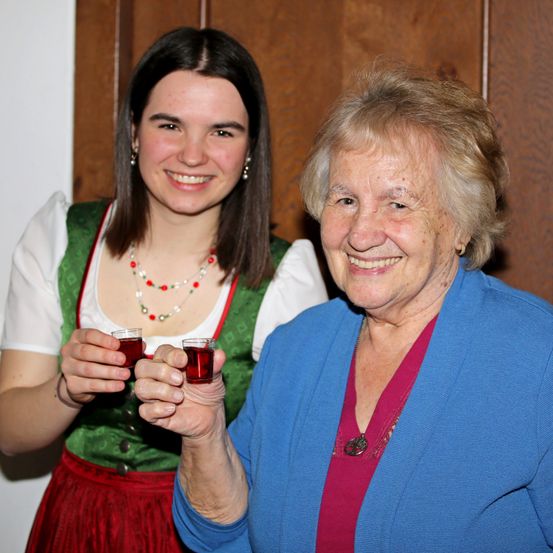 Zwei Frauen lächeln und halten kleine Gläser mit roter Flüssigkeit in einem Raum mit einer Holzwand. Die Frau links trägt ein grün-weißes Kleid mit einem roten Rock. Die Frau rechts trägt ein blaues Oberteil und eine Halskette.