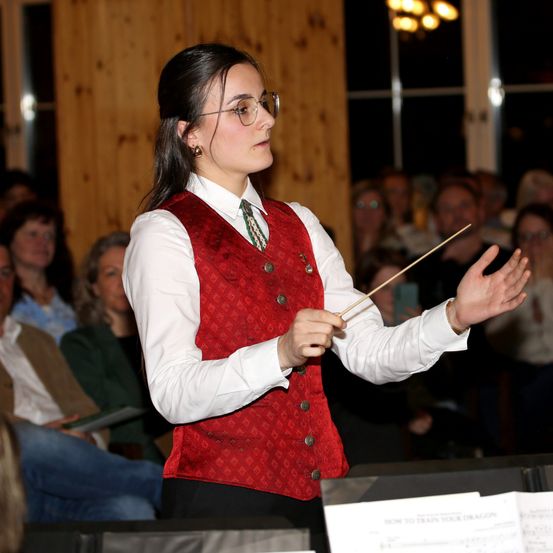 Eine junge Frau dirigiert ein Orchester mit einem Taktstock, trägt eine rote Weste mit einem weißen Hemd und steht vor einem Publikum.