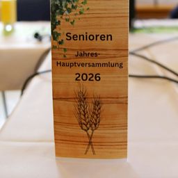 Eine hölzerne Karte mit der Aufschrift 'Senioren Jahres-Hauptversammlung 2026' und einer Weizen-Illustration auf einem Tisch.