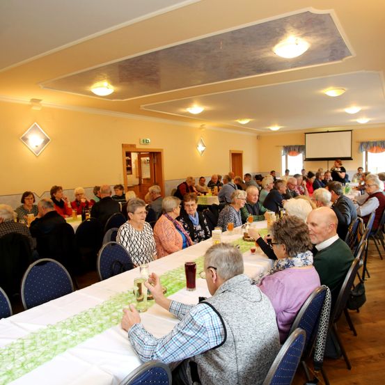 Eine große Gruppe älterer Menschen sitzt an langen Tischen in einem Saal. Die Tische sind mit weißen Tischdecken und grünen Tischläufern bedeckt. Mehrere Personen trinken, und eine Person hält ein Glas Wein. Der Raum ist durch Deckenleuchten beleuchtet.