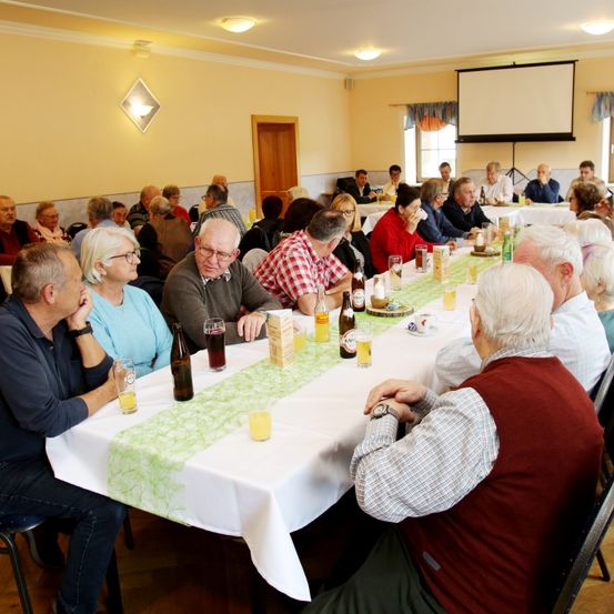 Eine Gruppe älterer Menschen sitzt um einen Tisch mit Getränken und Essen herum, möglicherweise bei einer Versammlung oder einem Treffen. Eine Projektionsleinwand ist an der Wand sichtbar.