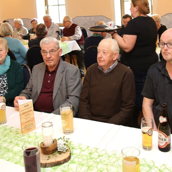 Mehrere ältere Personen sitzen um einen Tisch in einem Speisebereich. Sie schauen in die Kamera. Gläser mit Bier und Wein stehen auf dem Tisch.