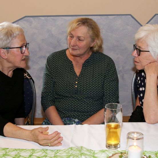 Drei ältere Frauen sitzen um einen Tisch herum, möglicherweise im Gespräch. Eine trägt eine Brille, eine andere einen Ring. Ein Glas Bier, eine Kerze und ein kleiner Baumstumpf stehen auf dem Tisch. Sie befinden sich in einem Raum mit einer gemusterten Wand.