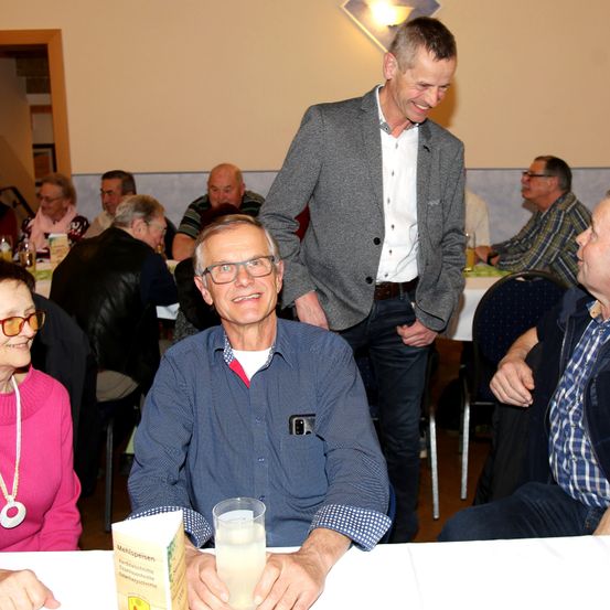 Eine Gruppe von Menschen sitzt an einem Tisch, ein Mann in einem blauen Hemd hält ein Glas. Ein anderer Mann steht hinter ihm und lächelt, während andere sitzen, einige mit Brillen und Jacken.