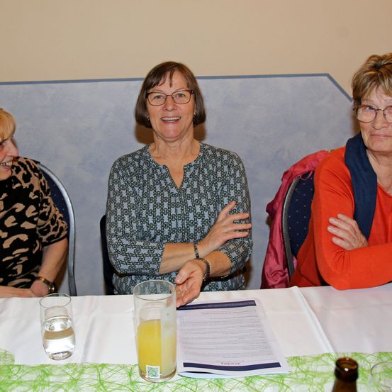 Drei Frauen sitzen um einen Tisch herum, lächeln und scheinen zu plaudern. Eine trägt eine Brille und ein gemustertes Hemd. Zwei Getränke und ein Dokument sind auf dem Tisch.